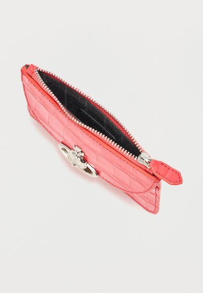Μικρό τσαντάκι από δέρμα με υφή κροκοδείλου σε κοραλί ροζ, με ασημένιο φερμουάρ και οβάλ μεταλλικό έμβλημα σε μονόχρωμο φόντο.