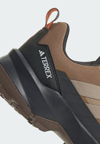 Brązowe i czarne buty sportowe z teksturowaną cholewką, z pomarańczową pętlą do ściągania i logo TERREX. Kontrastowe elementy wzornicze po bokach.