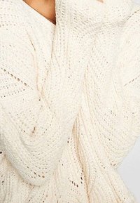 Pull en tricot couleur crème avec un motif en torsades texturé. Les manches longues sont amples, avec des points visibles et un aspect douillet.