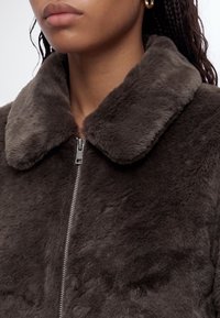 Femme portant une veste en fausse fourrure pelucheuse marron foncé avec un large col et une fermeture éclair argentée, affichée des épaules au menton.