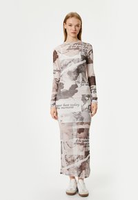 Pitkä, naisellinen mekko, joka on valmistettu läpinäkyvästä kankaasta, jossa on beige ja ruskea graafinen kuvio, jossa on tekstiä ja klassisia taideteemaja. Yhdistetty valkoisiin lenkkareihin.