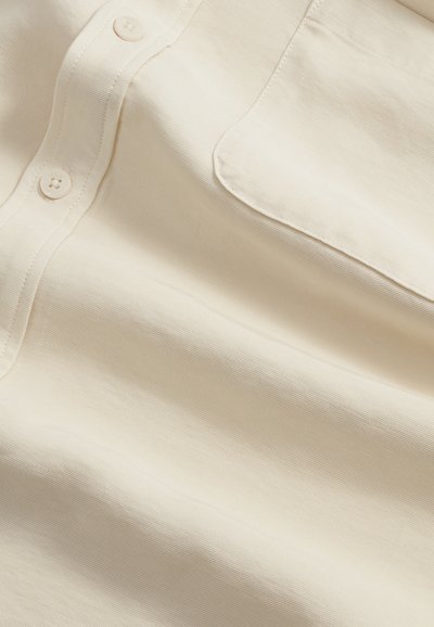 Tissu beige texturé avec une patte de boutonnage et une poche avant unique, montrant des coutures détaillées et le tissage du tissu.