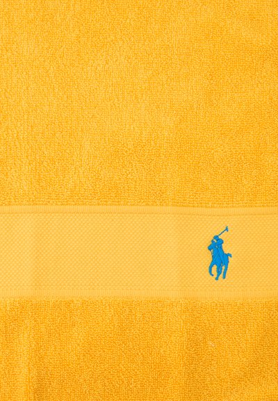 Κίτρινη πετσέτα με υφή, με οριζόντια ταινία που φέρει μικρό μπλε κεντημένο λογότυπο παίκτη πόλο πάνω σε άλογο.