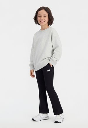 Uśmiechnięte dziecko z krótkimi kręconymi włosami, ubrane w jasnoszarą bluzę, czarne spodnie rozszerzane z logo oraz białe trampki, stojące na białym tle.