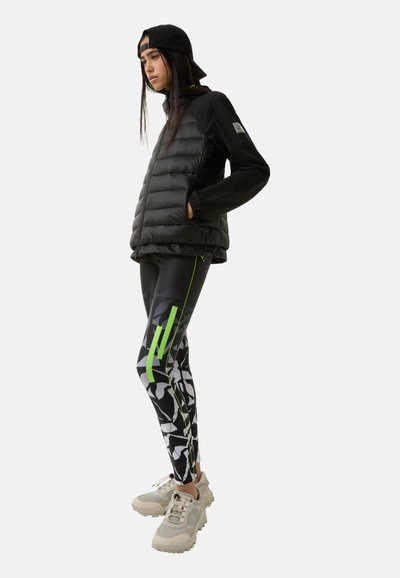 Sort pufferjakke med et elegant design, parret med mønstrede sorte leggings med grønne accenter og kraftige beige sneakers.