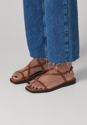 Piedi che indossano sandali piatti marroni con cinturini, smalto nero e jeans blu corti con orlo sfrangiato su un pavimento grigio chiaro.