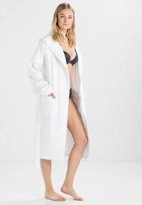 Witte badjas van zacht, gestructureerd stof, met capuchon, lange mouwen en voorzakken, gedragen over een zwarte beha en slipje.