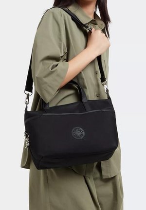 Persoon in een olijfgroen knopfouthemd die een zwarte schoudertas vasthoudt met twee handvatten en een verstelbare strap.