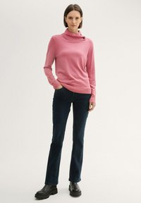 Rosa Pullover mit einem Schalkragen, ausgestattet mit zwei Knöpfen am Kragen, kombiniert mit dunkelblauen Jeans und schwarzen Plateauschuhen.