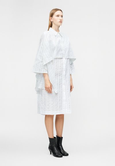 Γυναίκα που στέκεται, φορώντας λευκό χαλαρό φόρεμα μέχρι το γόνατο με κάθετες ρίγες και λεπτομέρεια πολλαπλών στρωμάτων στο μανδύα, συνδυασμένο με μαύρα μποτάκια αστραγάλου.