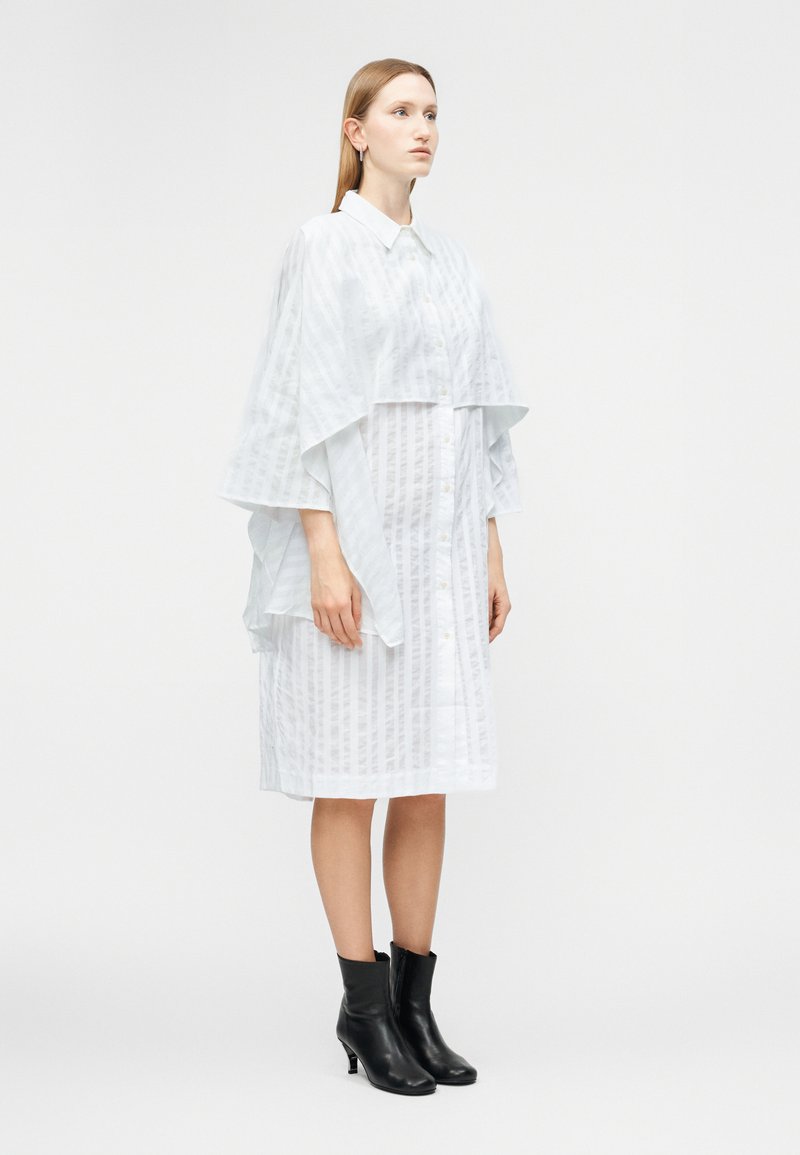 Femme debout, portant une robe blanche ample mi-longue à rayures verticales avec un détail de cape à couches, associée à des bottines noires.