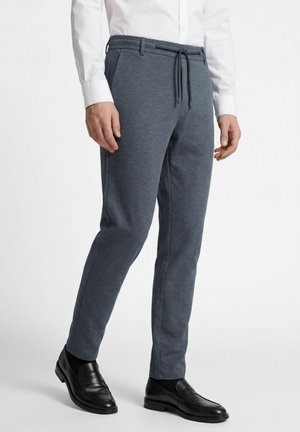 Man draagt donkergrijze slim-fit broek met trekkoord, wit hemd, zwarte sokken en zwarte leren loafers, staand tegen een witte achtergrond.