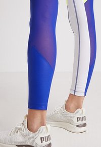 Bena iförda blå och vita träningsbyxor och vita PUMA stickade löparskor på en ljus bakgrund.