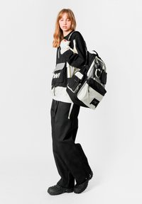 Mochila en blanco y negro con múltiples bolsillos, textura geométrica y correas ajustables, llevada con una chaqueta negra y pantalones negros holgados.