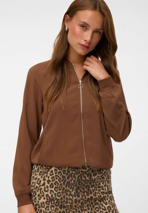 Woman with long brown hair wears a brown zip-up jacket and leopard print pants, touching her hair and looking slightly to the side.