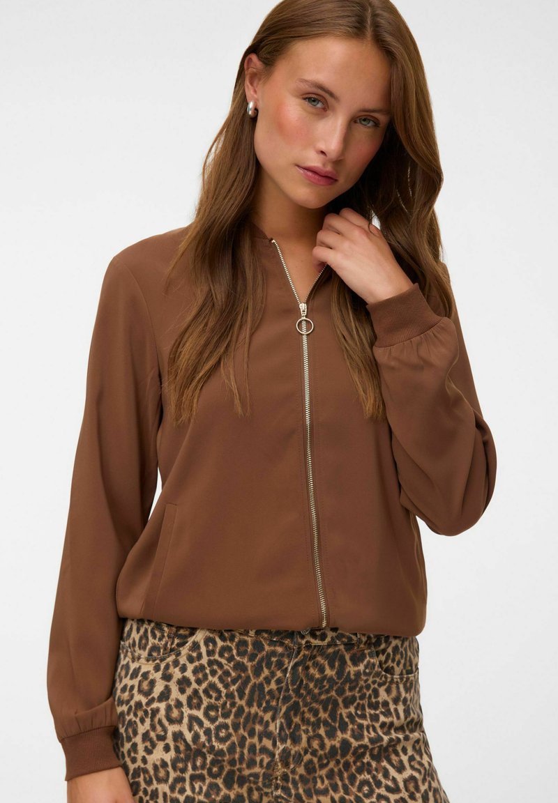 Femme aux longs cheveux bruns porte une veste marron zippée et un pantalon à imprimé léopard, elle touche ses cheveux et regarde légèrement sur le côté.