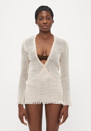 Cream-colored knitted wrap top with long sleeves, featuring a deep V-neck, frayed edges, and a lightweight, open weave texture.
