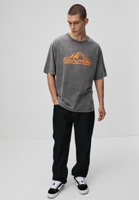 Młody mężczyzna z krótkimi włosami, ubrany w wyblakły szary, luźny t-shirt z pomarańczowym logo gór, czarne spodnie i czarno-białe trampki, stoi i patrzy w dół.