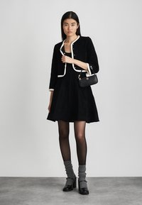 Svart sammetsklänning med en figursydd livstycke och utsvängd kjol, i kombination med en svart kort jacka med vit kant och en liten handväska.