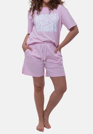 Donna con pantaloncini rosa e maglietta coordinata con la scritta "SUNDAYS", mani nelle tasche, in piedi a piedi nudi su sfondo bianco.