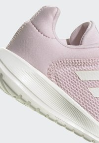Zapatilla deportiva rosa claro con parte superior de malla, cuello acolchado, suela blanca con surcos de tracción y tres rayas blancas en el costado.