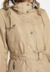Beige waterdichte jas met een trekkoord capuchon, zilveren knopen, een verstelbare taille en een ritszak aan de zijkant. Glad textuur van de stof.