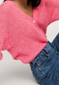 Vaaleanpunainen neulottu neuletakki, jossa on V-pääntie ja kolme nappia, yhdistettynä sinisiin korkeavyötäröisiin farkkuihin. Ominaisuuksiin kuuluu löysät hihat ja teksturoitu kuosi.