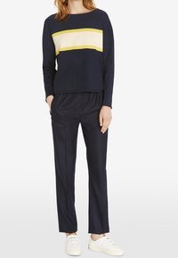 Maglia a maniche lunghe blu navy con motivo a righe crema e giallo, abbinata a pantaloni blu navy. Le sneakers sono bianche. Vestibilità rilassata, texture liscia.