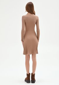 Geripptes, knielanges Kleid in Hellbraun mit langen Ärmeln. Eng anliegender Oberteil geht in einen ausgestellten Rock über. Kombiniert mit braunen Stiefeletten.