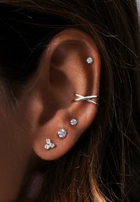 Pendientes de plata con diversos diseños: dos gemas transparentes engastadas, un racimo triangular y un aro entrelazado, sobre un tono de piel natural.