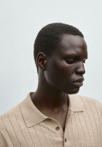 Beige strikket polo med lodrette striber, forsynet med krave og to knapper. Modellen har tætklippet mørkt hår og en glat teint.