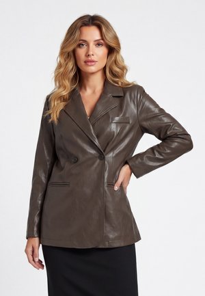 Vrouw met golvend blond haar die een donkerbruine leren dubbelgeknoopte blazer en een zwarte rok draagt, staand met één hand op haar heup.