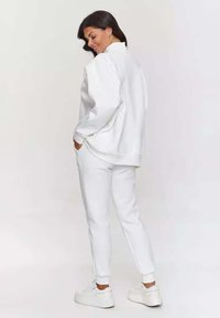 Biała bluza i dresy wykonane z miękkiego materiału. Luźny krój z opadniętymi ramionami, kieszeniami po bokach i zwężanymi mankietami. Białe trampki.