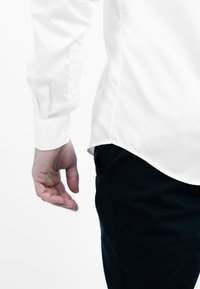 Witte long-sleeve shirt met knoopmanchetten, gecombineerd met donkere broek. Het shirt heeft een gebogen zoom en een gladde textuur.