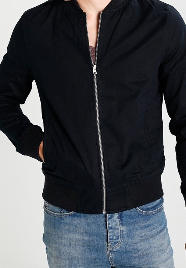 Person wearing a black zip-up jacket with hands in pockets and light blue jeans, shown from shoulders to hips.