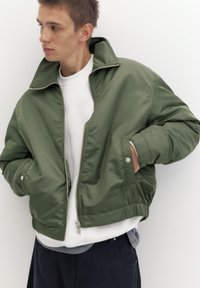 Jeune homme portant une veste bomber vert olive non zippée sur un sweat blanc, les mains dans les poches, regardant vers sa gauche.