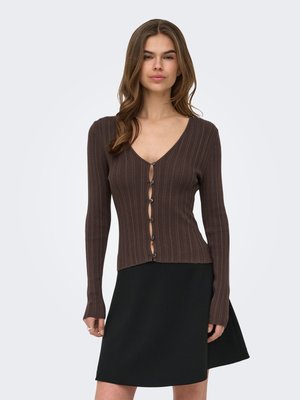 Kvinde iført en brun ribbet cardigan med knapper og et sort A-formet skørt, stående mod en ensfarvet lys baggrund.