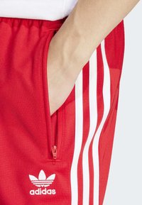Piros sportnadrág fehér csíkokkal, cipzáras zsebbel és fehér hímzett Adidas logóval. Simaság érzésű anyag.
