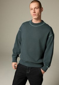 Tmavozelená mikina vyrobená z mäkkého materiálu, s okrúhlym výstrihom, rebrovanými manžetami a detailmi šitia. Skombinovaná s tmavými džínsami.