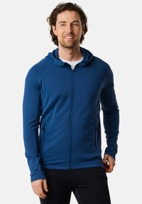 Felpa blu con zip realizzata in tessuto texturizzato, dal design aderente. Include un cappuccio e due tasche laterali, indossata sopra una maglietta bianca.