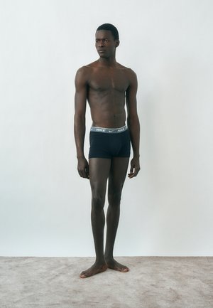 Férfi modell áll mezítláb szőnyegen, fekete Nike boxernadrágot viselve, egyszínű fehér háttér előtt.