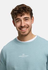 Ljusblå sweatshirt med rund hals, tillverkad av bomull, med en vit logga i mitten. Mjuk textur, avslappnad passform.