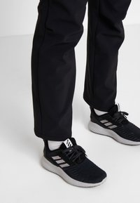 Svarta sportskor med texturerad mesh-övre, silverränder och vit sula, ihopparade med mörka byxor mot en neutral bakgrund.