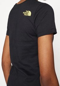 Černé bavlněné tričko s krátkým rukávem a kulatým výstřihem, na zádech žluté logo The North Face; hladká textura.