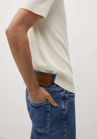 Persona vistiendo una camisa blanca de manga corta y jeans de mezclilla azules, con la mano casualmente en el bolsillo trasero, contra un fondo liso.