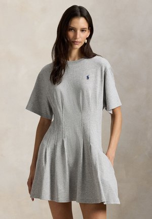 Femme aux longs cheveux bruns portant une robe grise à manches courtes avec des poches latérales et un petit logo brodé sur la poitrine, debout devant un fond neutre.