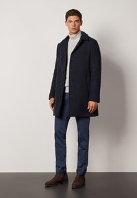 Marinblå ullkappa med en strukturerad krage, framknäppning, sidfickor och en skräddarsydd passform, kombinerad med en grå polo och marinblå byxor.