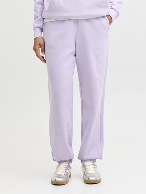 Persona con pantalones de chándal morados claros y sudadera a juego con las manos en los bolsillos, combinados con zapatillas plateadas brillantes con suelas beige.