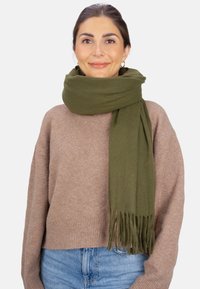 Écharpe verte en tissu doux, avec des bords à franges, portée sur un pull marron clair, sur fond blanc uni.