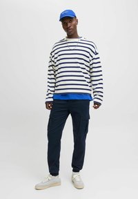 Langärmliges gestreiftes Hemd in Weiß und Marineblau, darüber ein blaues Hemd getragen. Marineblaue Cargohosen mit Taschen. Graue Sneakers mit beigen Sohlen. Blaue Kappe.